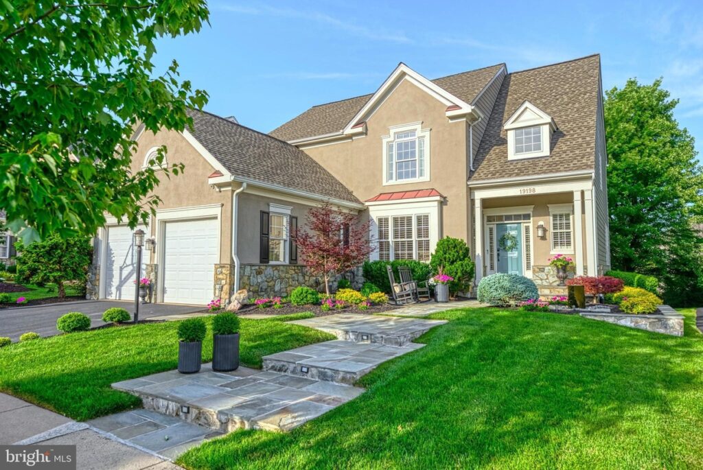 This home is part of the April Loudoun County Real Estate Update, which provides information on homes for sale in Loudoun County.
An inviting two-story single-family home located at 19198 CHARTIER DRIVE, LEESBURG, VA 20176, featuring a blend of stone and stucco facade with contrasting shutters. A well-manicured lawn with vibrant flower beds complements the home's warm beige exterior. The entrance boasts a cozy porch area with a rocking chair, perfect for enjoying the serene suburban neighborhood