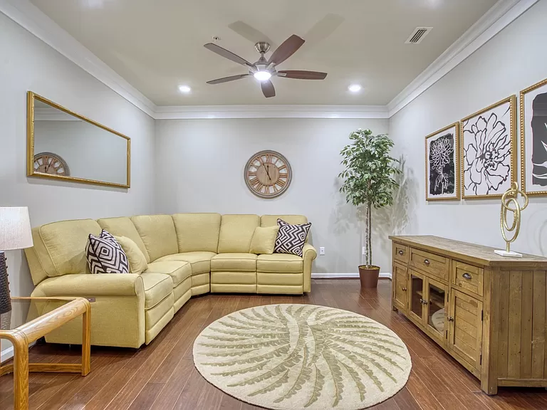 The image shows a stylishly decorated living room. It features a large, soft yellow sectional sofa that comfortably fills a corner of the room, complemented by patterned throw pillows. The flooring is a rich, dark hardwood that contrasts nicely with the pale walls and ceiling. There's a decorative circular rug in the center of the room, along with a rustic wooden sideboard against one wall. Artwork with floral motifs in ornate frames and a large wall clock with Roman numerals enhance the room's elegant ambiance. A ceiling fan above adds a practical touch while a potted plant brings a bit of nature indoors, completing the cozy, welcoming space.