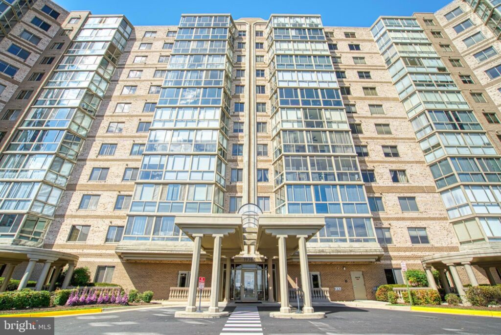 This home is part of the April Loudoun County Real Estate Update, which provides information on homes for sale in Loudoun County.
The grand entrance of a high-rise condominium building at 19365 Cypress Ridge Terrace Unit 611, Leesburg, VA 20176, with an elegant portico supported by columns. The building's facade is adorned with numerous glass balconies and windows, reflecting a sunny blue sky, and is accented with tasteful landscaping.